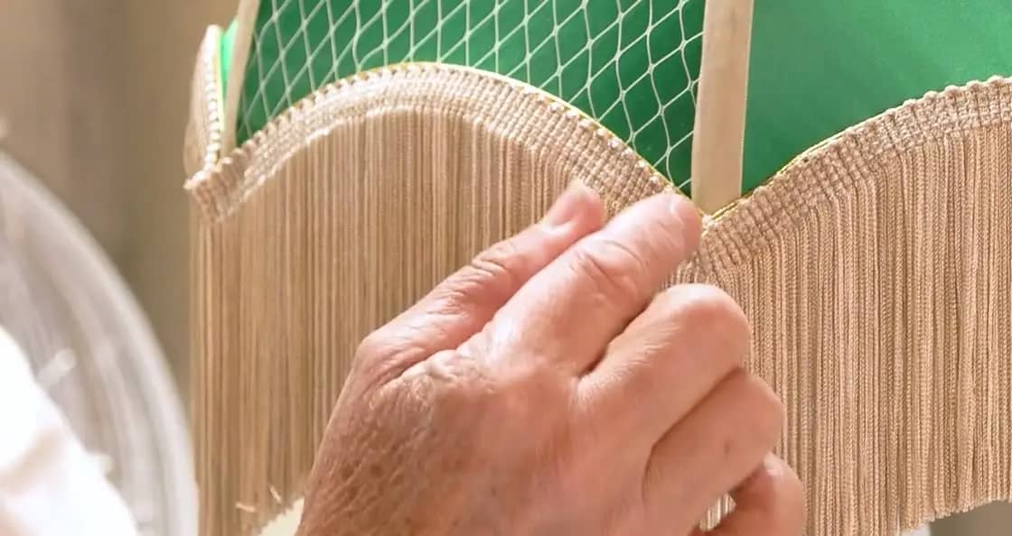 La main de Sylvie Lemarchand dans la frange d’un abat-jour dôme en satin vert, à l’atelier